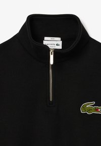 Sudadera negra con cremallera y cuello alto, con una cremallera plateada y un logo de cocodrilo bordado en verde en el pecho. Corte holgado.