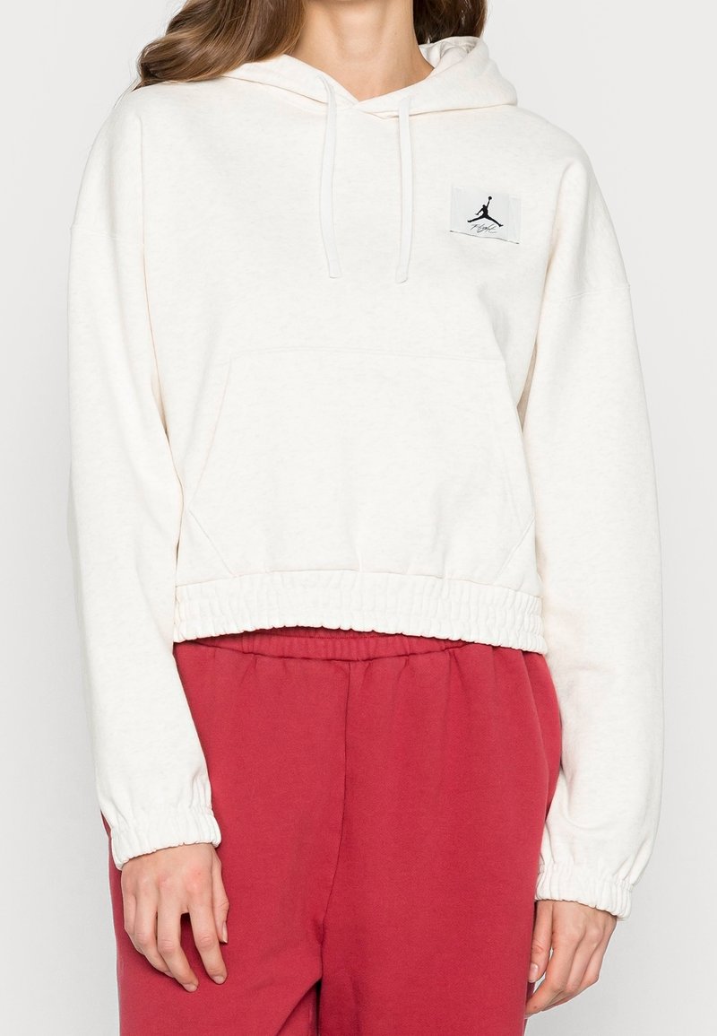 Vit kort hoodie gjord av mjukt tyg med ribbad nederkant, med en framficka och svart Jordan-logotyp på bröstet.