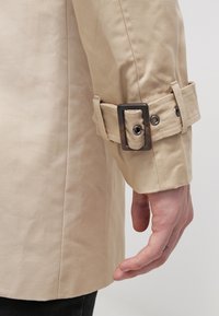 Béžový trenčkot s hladkou texturou, s přezkou a páskem na manžetě. Šití je viditelné podél švu rukávu.