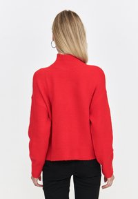 Czerwona dziergana sweter z wysokim kołnierzem, oversize'owy krój, opadające ramiona i ściągacz na dole. Miękka tekstura i skrócony fason.