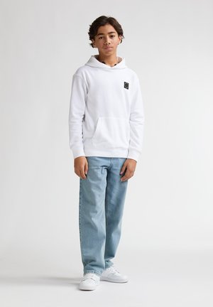 Hanorac alb cu glugă și buzunar cangur, purtat cu blugi deschis la culoare și adidași albi. Are un mic logo negru pe piept.