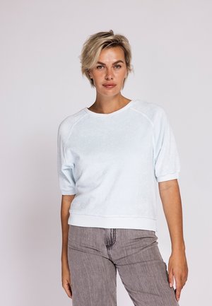 Frau mit kurzen blonden Haaren, die einen hellblauen kurzärmligen Pullover und graue Jeans trägt, steht vor einem einfachen hellen Hintergrund.