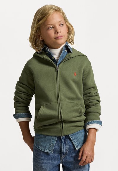 Olivno zelena zip-up kapuca iz mehkega materiala s sprednjim žepom in rdečim logotipom. Narejena je nad denim srajco s zvitimi rokavi in modrimi kavbojkami.