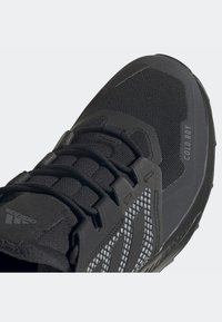Gros plan d'une basket Adidas noire avec dessus en mesh, lacets noirs, et texte "COLD.RDY" sur le côté près de la zone des orteils.