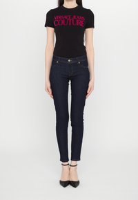 Svart t-shirt med kort ärm med "VERSACE JEANS COUTURE" i knallrosa. Sammanpassade mörkblå jeans i slim fit, tillsammans med svarta spetsiga klackar.