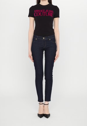 Czarna koszulka z krótkim rękawem z napisem "VERSACE JEANS COUTURE" w intensywnym różowym kolorze. Dopasowane, ciemnoniebieskie dżinsy o wąskim kroju, zestawione z czarnymi szpilkami.