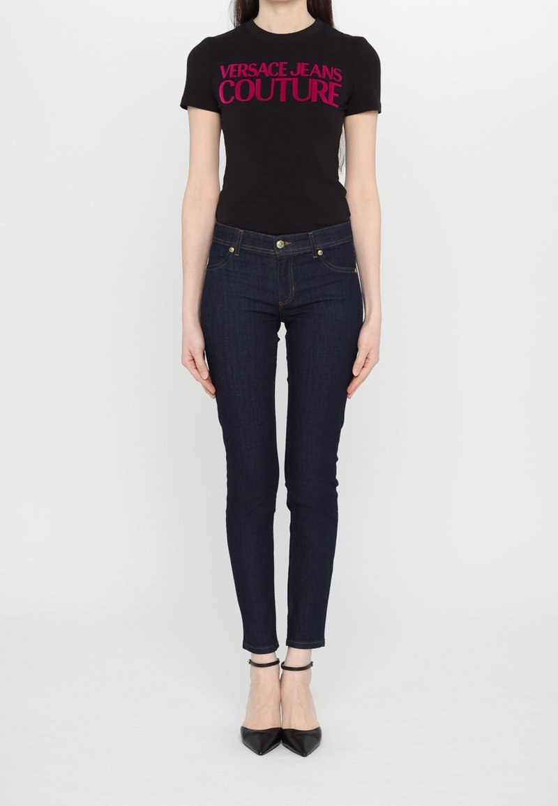 Svart t-shirt med kort ärm med "VERSACE JEANS COUTURE" i knallrosa. Sammanpassade mörkblå jeans i slim fit, tillsammans med svarta spetsiga klackar.