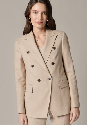 Kobieta w beżowej, dwurzędowej marynarce z ciemnymi guzikami i dopasowanych spodniach, stojąca na jednolitym neutralnym tle.