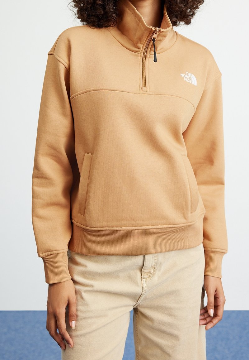 Personne portant un sweat North Face beige clair à fermeture quart-zip et un pantalon beige clair, debout avec une main vers le bas dans un environnement intérieur neutre.