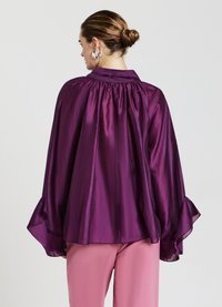 Blusa viola fluida con colletto alto, dettagli arricciati sul retro e ampie maniche a campana. Tessuto liscio, abbinata a pantaloni rosa chiaro.