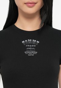 Juoda juostuota marškinėliai su trumpomis rankovėmis, puošti balta tekstine logotipu "TOMMY JEANS" ir vietos detalėmis. Klasikinis apvalus kaklas, prigludęs dizainas.