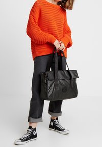 Sac fourre-tout noir en tissu texturé, à poignée plate et rabat supérieur, tenu par une personne portant un pull en maille orange et un jean noir aux revers retroussés.