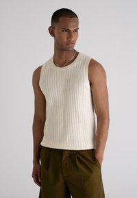 Ribbed sleeveless knit top in off-white, featuring a round neckline and subtle logo detail. Paired with dark olive green trousers.