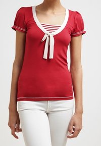 Mujer vistiendo una blusa roja de manga corta con ribetes blancos y un lazo sobre una camiseta de rayas rojas y blancas, combinada con pantalones blancos.
