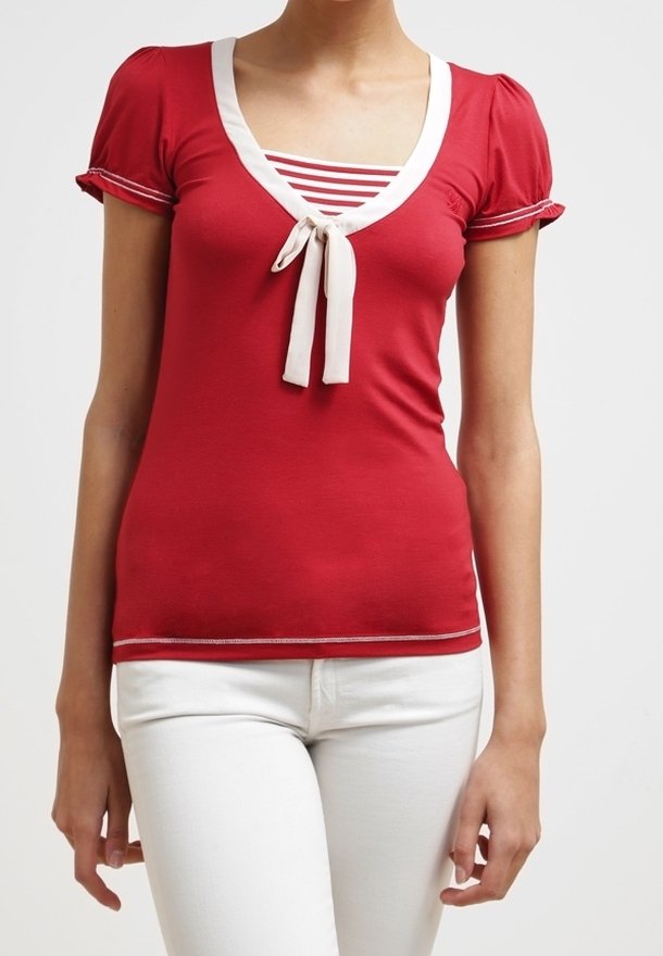 Mujer vistiendo una blusa roja de manga corta con ribetes blancos y un lazo sobre una camiseta de rayas rojas y blancas, combinada con pantalones blancos.