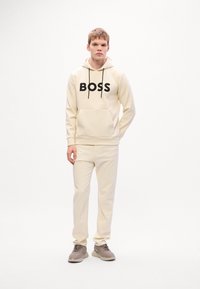 Jeune homme portant un sweat à capuche crème avec le texte "BOSS", un pantalon crème assorti et des baskets grises, se tenant devant un fond blanc uni.