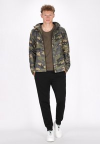 Kamuflagejakke i nuancer af grøn og brun, parret med en brun skjorte og sorte bukser. Hvide sneakers fuldender outfittet.