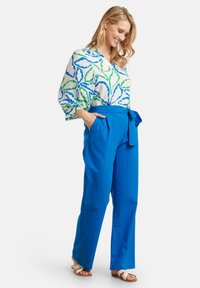 White blouse with blue and green leaf pattern, paired with blue wide-leg trousers featuring a tie waist and side pockets. Tan sandals.