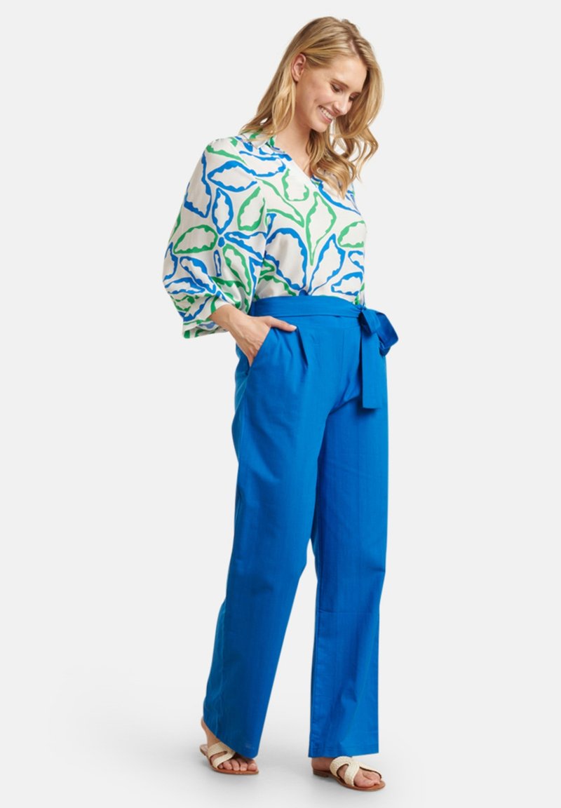 White blouse with blue and green leaf pattern, paired with blue wide-leg trousers featuring a tie waist and side pockets. Tan sandals.
