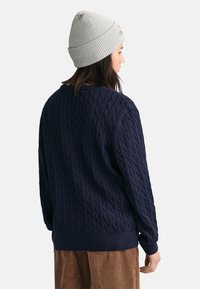 Marineblauer Kabelstrickpullover mit strukturiertem Muster und gerippten Bündchen, kombiniert mit einer hellgrauen Mütze und Cordhosen.