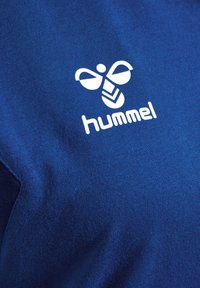 Blå stof t-shirt med et hvidt Hummel-logo. Materialet har en glat overflade med et subtilt ribbet mønster.