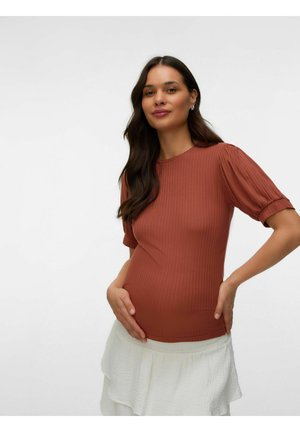 Donna incinta con lunghi capelli scuri che indossa un top color ruggine e una gonna bianca, che tiene delicatamente la pancia, sorridendo dolcemente.