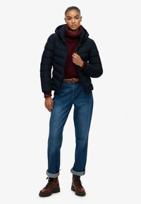 Marineblauwe pufferjas met een capuchon, gequilt ontwerp en ritssluiting. Gecombineerd met een bordeauxkleurige turtleneck, blauwe jeans en bruine veterschoenen.