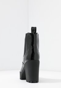 Botas de tobillo de charol negro con tacón robusto, paneles laterales elásticos y acabado brillante, que cuentan con costuras decorativas y una tira de agarre.