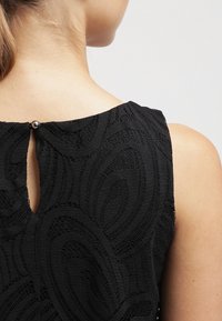 Gros plan sur le haut du dos et l’épaule d’une femme portant un haut sans manches noir à motif en dentelle avec une ouverture boutonnée au niveau de l’encolure.