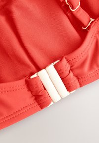 Piros bordázott szövet sima textúrával, fehér kapoccsal és elasztikus részletekkel a szélén. Közeli kép a dizájnról és a fémalkatrészekről.