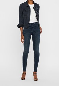 Chaqueta de mezclilla oscura con dos bolsillos en el pecho, combinada con jeans oscuros de cintura alta y corte ajustado, y tacones marrones de tiras sobre un fondo liso.