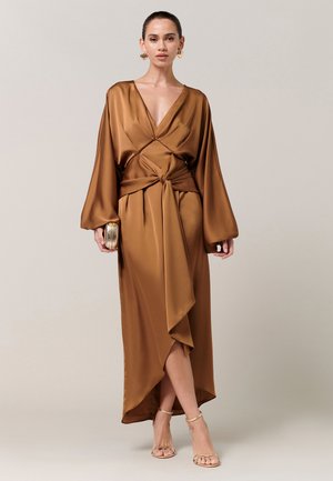 Donna che indossa un abito incrociato in raso bronzo con scollo a V profondo, maniche larghe, vita annodata, tiene in mano una piccola pochette metallica e indossa tacchi dorati.