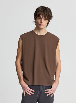 Giovane uomo con capelli castano ondulati, che indossa una maglietta marrone senza maniche e pantaloni scuri, in piedi con le mani vicino alle tasche contro uno sfondo neutro.