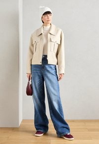 Chaqueta corta beige con dos bolsillos frontales, suéter claro debajo, jeans anchos azules y zapatillas burdeos. Gorro de punto blanco con pompón.