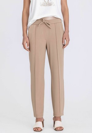 Beige broek met hoge taille en taps toelopende pijpen, voorzien van plooien aan de voorkant en een gestrikte tailleband, gedragen met een wit mouwloos topje en open teen hakken.