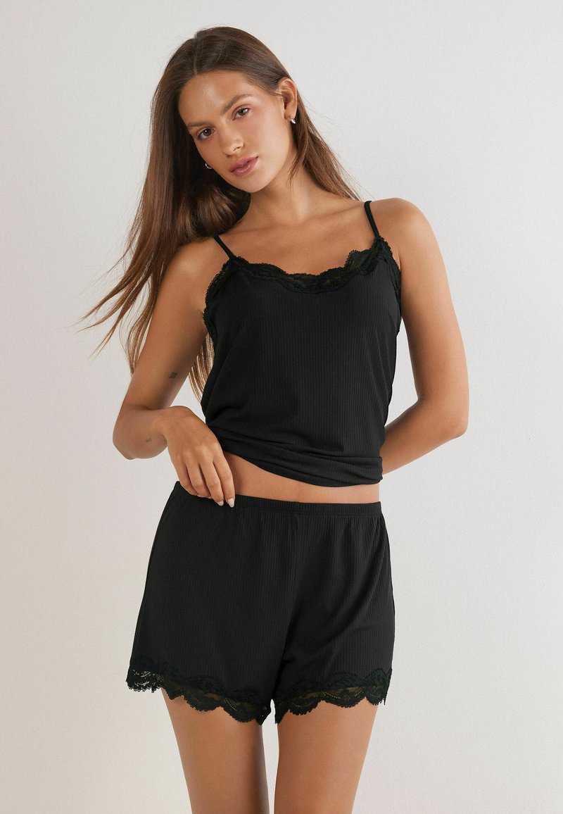 Vrouw in een zwarte kanten top met bijpassende shorts, staand tegen een effen witte achtergrond.