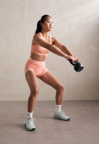 Kvinnelig idrettsutøver i en myk rosa sports-BH og shorts, som bruker en svart kettlebell. Hun har på seg mintgrønne joggesko og hvite sokker, på en lys bakgrunn.