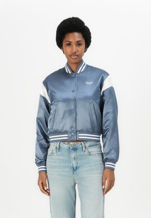 Giacca bomber in satin blu navy con strisce bianche, polsini a coste e un logo, caratterizzata da una silhouette corta e chiusura a bottoni a pressione.