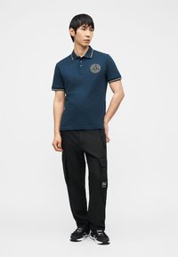 Άνδρας που φοράει μπλε κοντομάνικο πόλο με λογότυπο στο στήθος, μαύρο παντελόνι cargo και μαύρα αθλητικά παπούτσια, στέκεται με το ένα χέρι πίσω από την πλάτη.