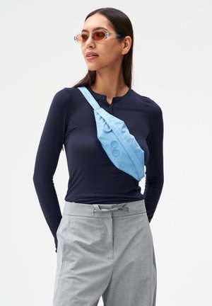 Frau trägt orangetönte Sonnenbrille, dunkelblaues Langarmshirt, hellgraue Hose und eine hellblaue Umhängetasche vor weißem Hintergrund.