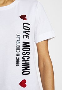 Vit bomulls t-shirt med svart tryckt text "LOVE MOSCHINO {ETABLERAD 2008}" och en röd hjärtaccent på vänster sida.