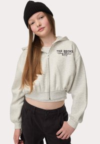 Grijze cropped hoodie met rits, voorzien van de tekst "THE BRONX" op de borst, gecombineerd met zwarte broek. Zachte stof en een ontspannen pasvorm.