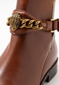 Botines de cuero marrón con un acento de cadena dorada y un cierre decorativo, textura suave y un tacón bajo cuadrado con detalles de tachuelas.