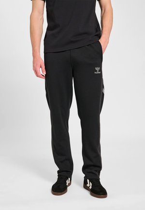 Mężczyzna ubrany w czarne spodnie dresowe Hummel i czarne trampki z białymi akcentami, stojący z rękami blisko kieszeni na szarym tle.