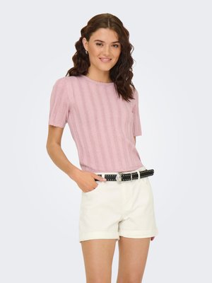 Jonge vrouw met golvend bruin haar, draagt een roze gebreide top met korte mouwen, witte korte broek en een zwarte gespikte riem, glimlacht met één hand in de zak.