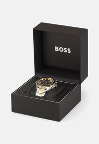 Reloj en tonos plateado y dorado con un dial negro, ubicado en una caja negra con el logo blanco "BOSS". Presenta una pulsera de metal y un bisel detallado.