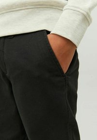 Handen i fickan på svarta byxor som bärs med en ljusbeige långärmad topp mot en enfärgad bakgrund.