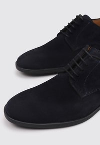 Chaussures en daim bleu marine avec une texture lisse, bout rond et système de laçage à six œillets. Semelle en caoutchouc avec un profil de traction minimal.