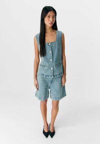 Gilet di jeans con scollo quadrato e bottoni frontali, abbinato a pantaloni corti in jeans coordinati. Entrambi di un blu chiaro con una leggera sfumatura. Tacchi a punta neri.