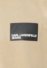 Beige puuvillapaita, jossa on musta suorakulmainen etiketti, jossa lukee "KARL LAGERFELD JEANS" valkoisella lihavoidulla tekstillä. Suorakulmainen, rento muotoilu.